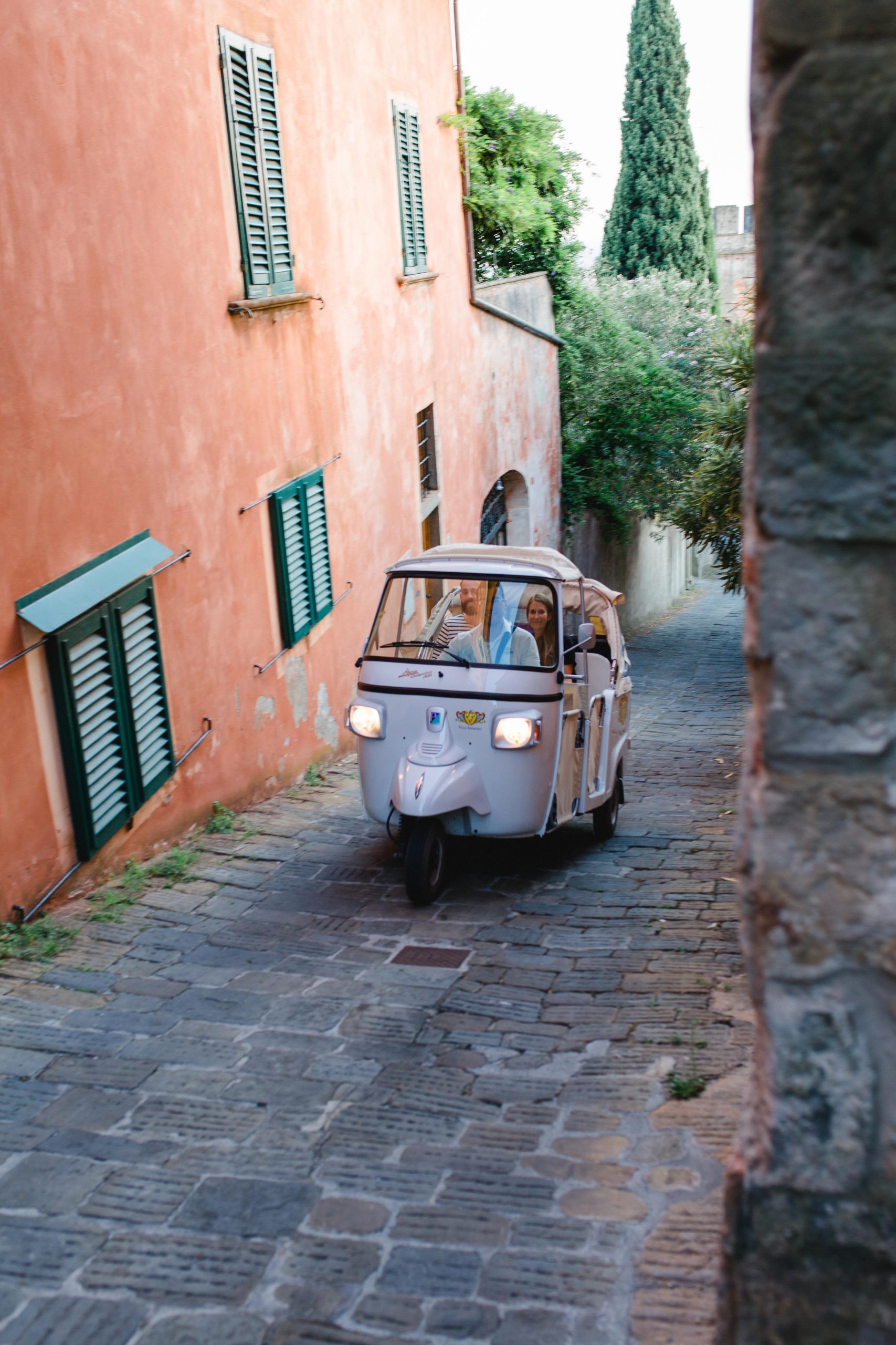 Small three-wheeled car driving up a narrow cobblestone street beside terracotta walls with green shutters and trees.